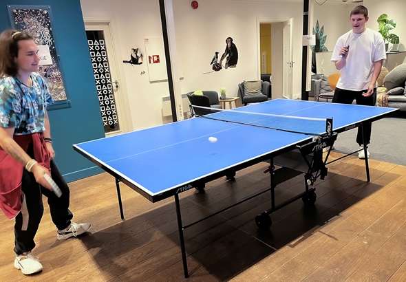 To personer går mot sykehuset på Røde Kors, med parkeringsplasser og moderne bygning i bakgrunnen. Godt vær og avslappet atmosfære.