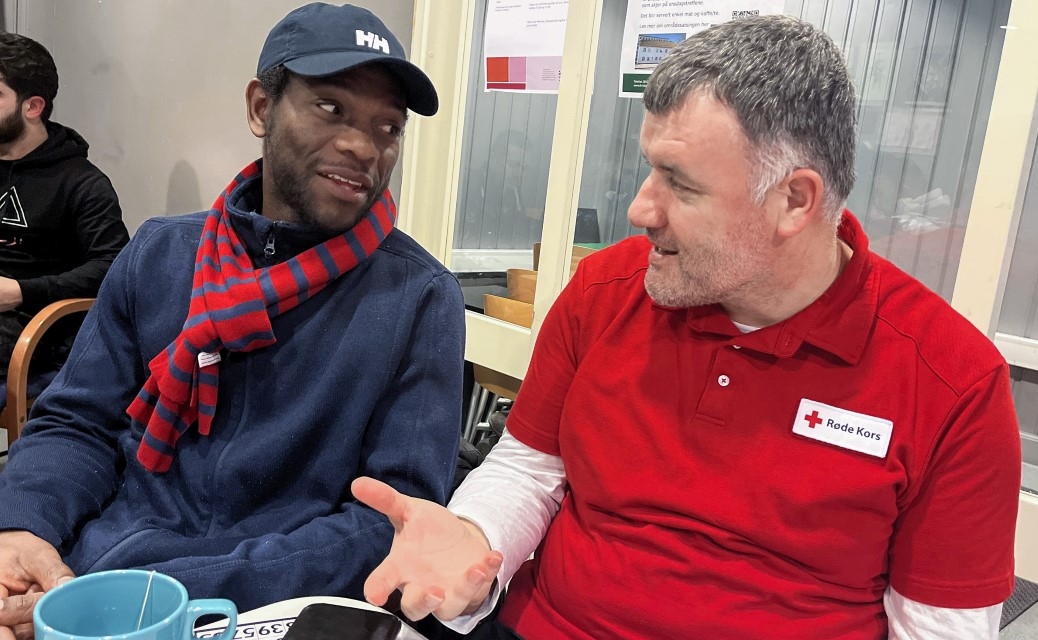 En mann i Røde Kors-skjorte sitter og snakker med en deltaker ved et bord. De ser på hverandre og er i samtale, med kaffekopper på bordet foran seg.