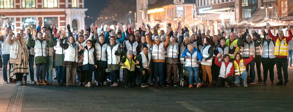 En stor gruppe Røde Kors-frivillige står samlet ute i en opplyst bygate om kvelden. De har på seg refleksvester og løfter hendene i været mens de smiler mot kameraet. Gruppen består av mennesker i ulik alder og bakgrunn, inkludert en person i rullestol. Lysene fra butikker og bygninger skaper en varm stemning bak dem.