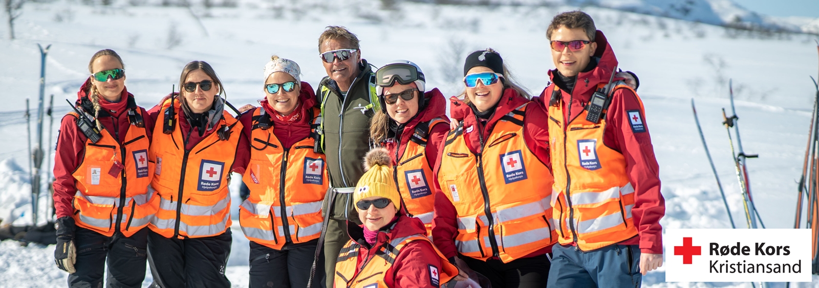 En gruppe Røde Kors-frivillige står samlet ute i et snødekt fjellområde, alle iført røde jakker og oransje refleksvester. De smiler mot kameraet i solskinnet, og en liten hund i sele står foran gruppen. I bakgrunnen ses skiutstyr og en person med lue som ser mot gruppen.