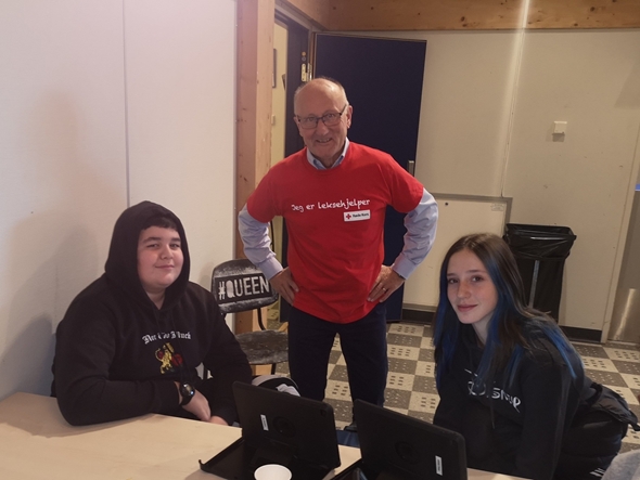 En mann i rød leksehjelper-skjorte står sammen med to ungdommer som sitter