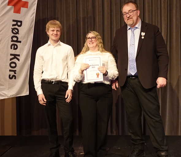 To personer mottar pris for årets forening i Hedmark Røde Kors. De står på en scene sammen med en mann.