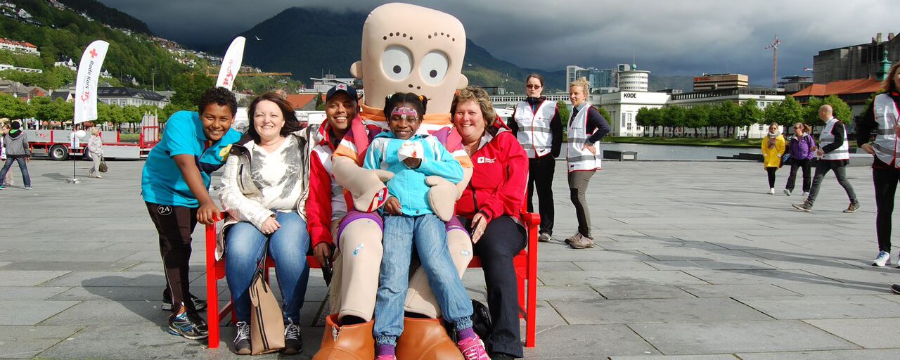 Barn og voksne sitter på rød benk på en offentlig plass i Bergen