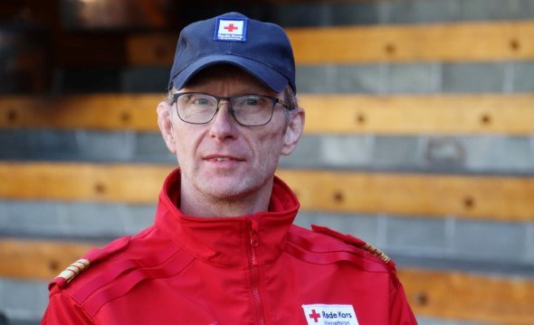 En mann med rød uniformsjakke og blå lue med kikker rett inn i kamera. Han har på seg briller.