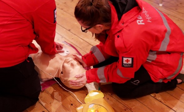 Bildet viser to personer med rød uniform fra Rede Kors som viser hjerte- og lungeredning på en dukke.