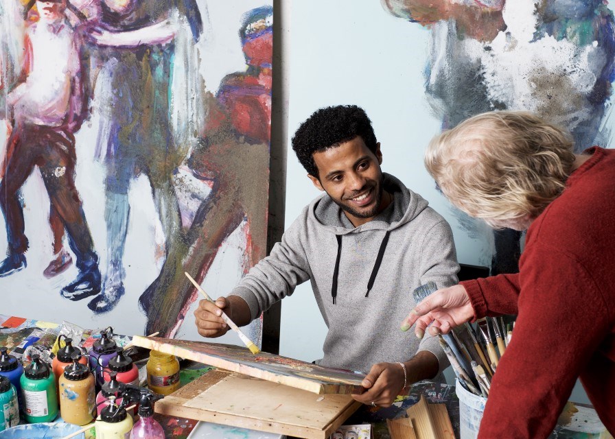 En ung mann med mørk hud sitter i et atelier og maler mens han snakker med og smiler til en eldre person som forklarer ham noe.