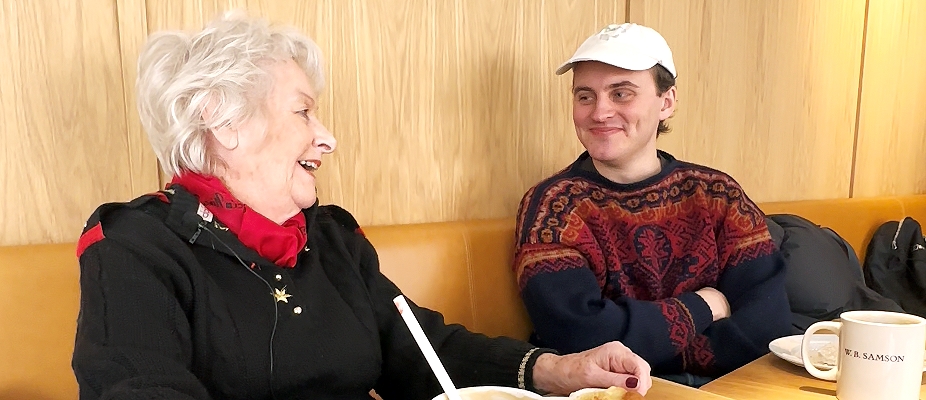 Besøksvennepar Inger (83) og Endre (24) på café. De ler. 