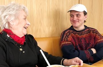 Besøksvennepar Inger (83) og Endre (24) på café. De ler. 