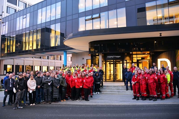 En stor gruppe mennesker, kledd i ulike uniformer, står samlet foran en bygning med store glassfasader og lysende vinduer.