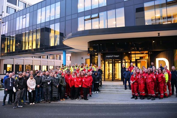 En stor gruppe mennesker, kledd i ulike uniformer, står samlet foran en bygning med store glassfasader og lysende vinduer.