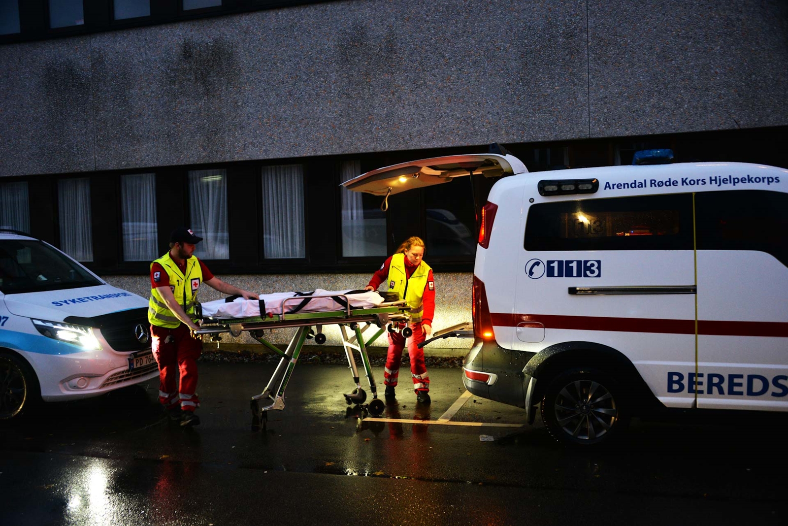To personer i Røde Kors-uniform håndterer en båre foran en beredskapsambulanse. Det er mørkt ute med våt asfalt som reflekterer lys.