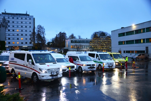 Flere beredskapsambulanser og ambulanser står parkert i skumringen. Bilene har synlige logoer fra Røde Kors og nødetater. I bakgrunnen er det bygninger med julelys i trærne, og himmelen er blågrå.