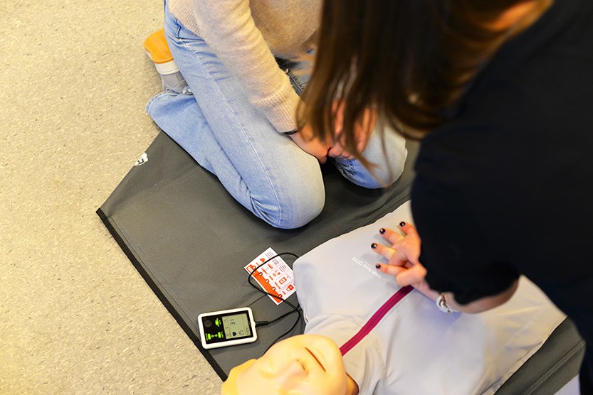 To personer utfører hjerte-lungeredning på en treningsdukke som ligger på en matte. Ved siden av dukken ligger en elektronisk enhet som gir tilbakemelding på kvaliteten av kompresjonene.