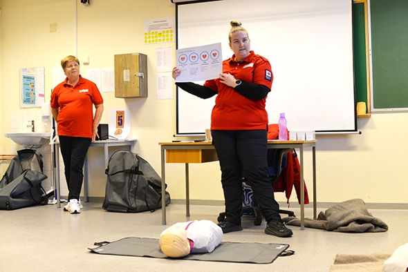 En person holder opp et informasjonsark med illustrasjoner av HLR-trinn foran en projektorskjerm, mens en treningsdukke ligger på en matte på gulvet. I bakgrunnen står en annen person i rødt kursantantrekk.