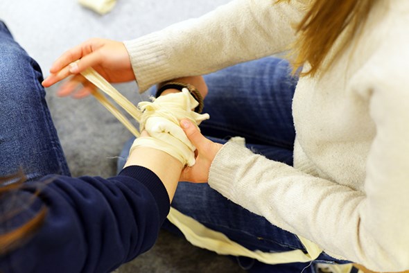 To personer sitter på gulvet mens den ene legger en trykkbandasje rundt håndleddet til den andre. Bandasjen er hvit og surres stramt for å simulere behandling av skade.