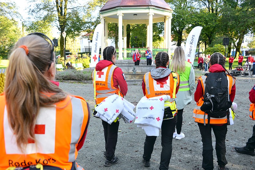 Flere personer i Røde Kors-refleksvester går mot en paviljong i en park, bærende på tepper med Røde Kors-logo. I bakgrunnen står andre frivillige og bannere med Røde Kors-logo, omgitt av trær og grøntområde.