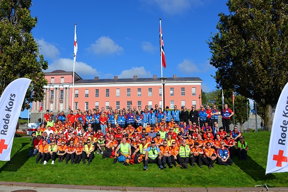 Gruppebilde av mange Røde Kors-frivillige i fargerike uniformer og refleksvester, sittende og stående på en gressplen foran en stor rosa bygning med flaggstenger og norske flagg, omgitt av trær og bannere med Røde Kors-logo.