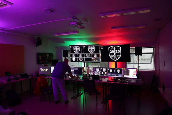 Rom med mange monitorer som viser logoen «NM 2025», fargerike lys i taket, høyttalere og teknisk utstyr på bordene, mens en person justerer oppsettet foran skjermene.