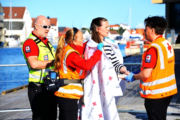 Tre personer i Røde Kors-uniform og refleksvester hjelper en person som er innpakket i et teppe, ved en brygge med vann, båter og hus i bakgrunnen.
