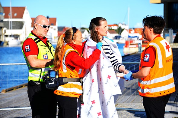 Tre personer i Røde Kors-uniform og refleksvester hjelper en person som er innpakket i et teppe, ved en brygge med vann, båter og hus i bakgrunnen.