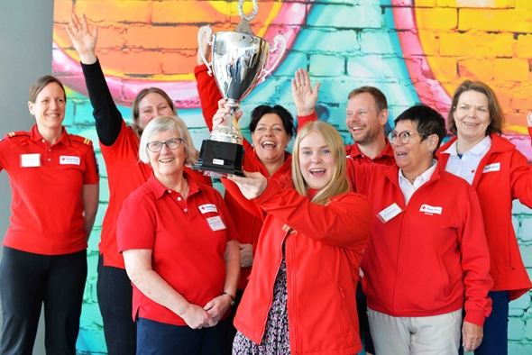Bilde av åtte smilende mennesker i Røde Kors-klær som løfter pokal.