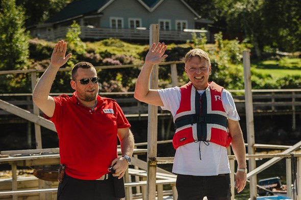 To menn som vinkar, ein i Røde Kors t-skjorte og ein i redningsvest med norsk flagg