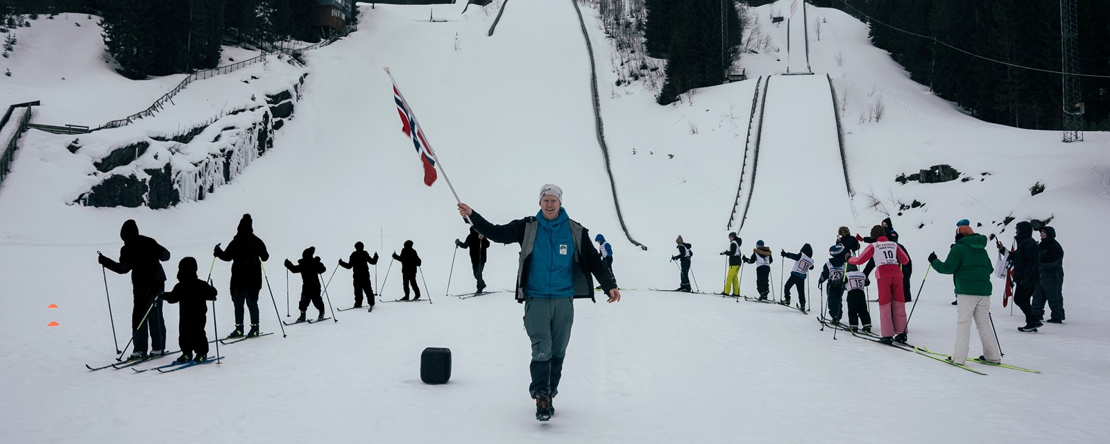 Barn og vaksne på ski i sirkel rundt mann med norsk flagg