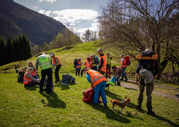 Ei gruppe frivillige frå Røde Kors sit i graset og et lunsj i sola. Dei har på seg turklede og refleksvestar, og stemninga er avslappa og god. Skog og blå himmel i bakgrunnen.