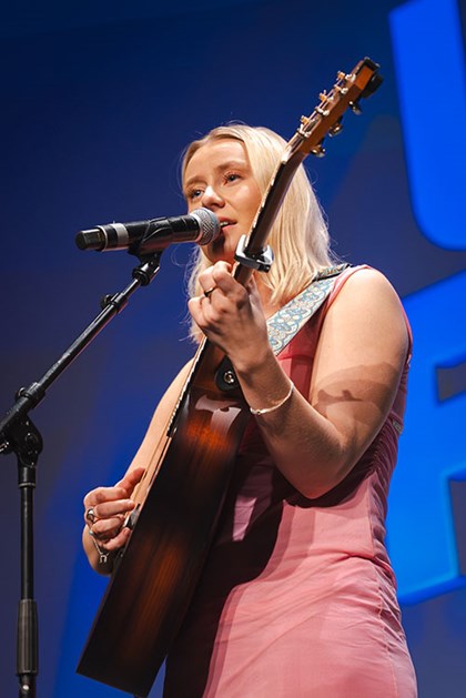 Ei kvinne syng på scena, med mikrofon og gitar