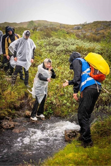Ein mann i Røde Kors vest strekkjer ut ei hand til ein deltakar i det dei hoppar over ein bekk.