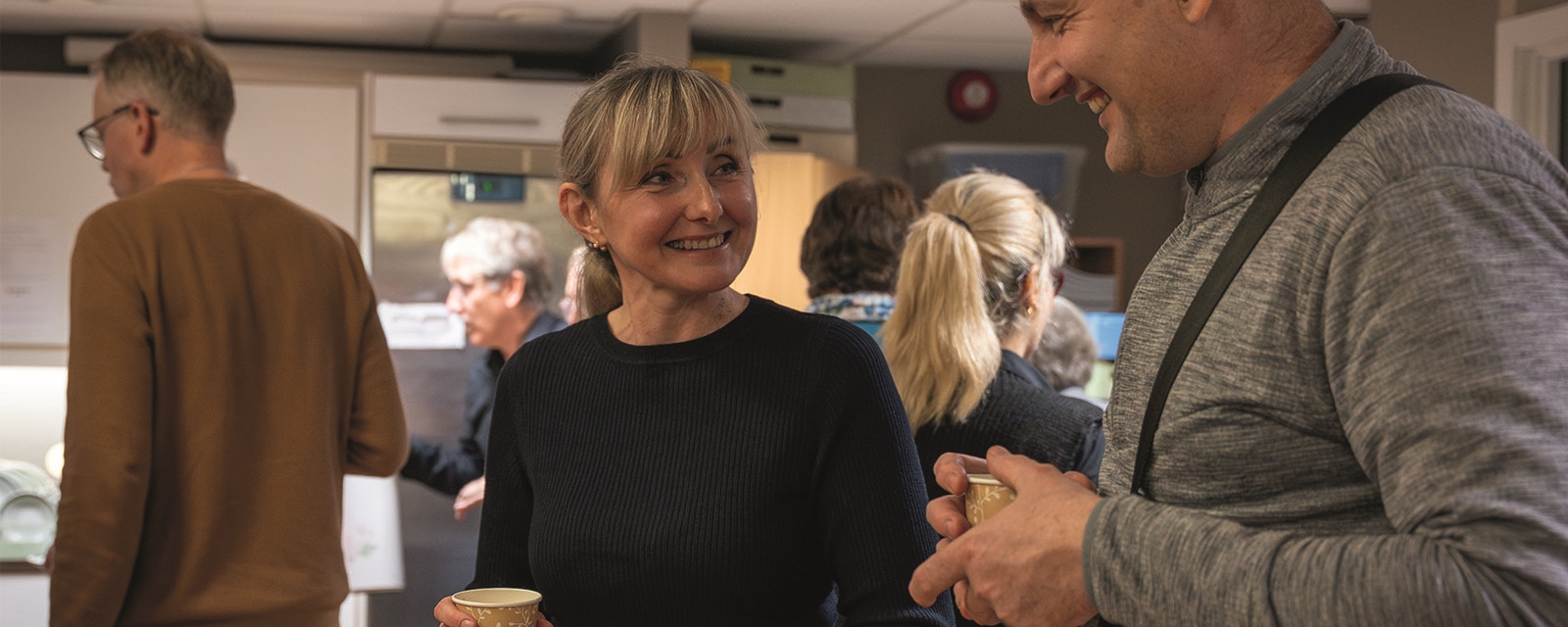 Ei kvinne held ein kaffekopp og smiler mot ein mann som ler.