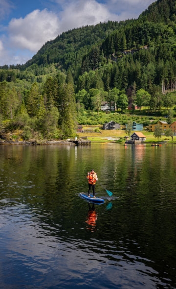 Jente står på paddleboard på stille vatn, omgitt av grøn natur ved Haugland.