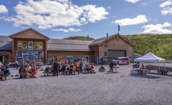 Gruppe menneske sit og står utanfor Hodlekve skisenter på ein solrik dag. Nokre et, nokre snakkar, og fleire kviler etter fjelltur. Røde Kors-telt og fjell i bakgrunnen.