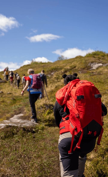 Fjellvandrarar går oppover ein sti i terrenget, med ryggsekkar og fjellklede. Forgrunnen viser ein person med raud sekk, og landskapet er grønt og ope under blå himmel.