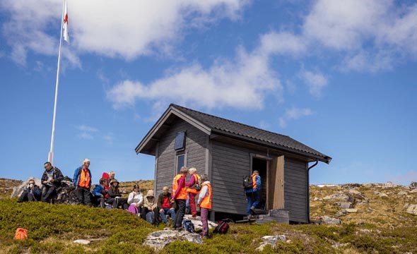 Ei gruppe menneske sit og står utanfor ei lita, mørkgrå hytte med Røde Kors-flagg ved Britastein. Det er sol, og landskapet rundt er fjell og lyng.