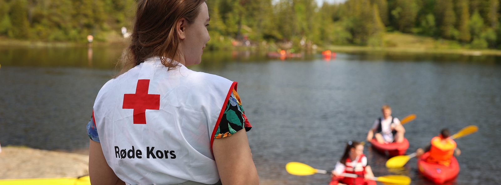 Frivillig i vest med Røde Kors-logo speider utover sjøen hvor flere deltagere padler kajakk.