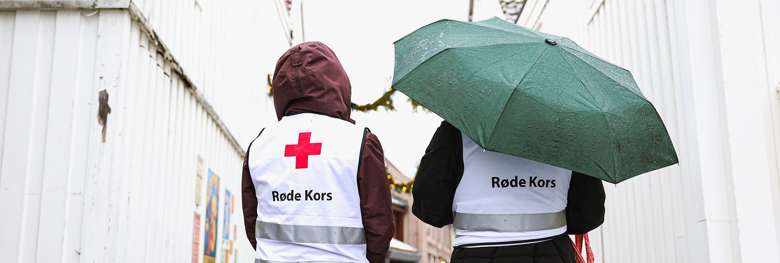 Ryggen til to med Røde Kors vest som står ute i regnet med en paraply.