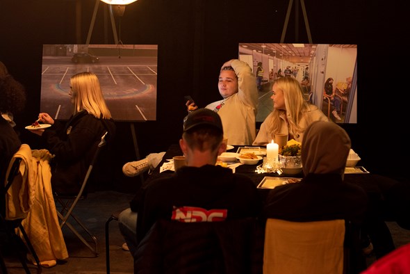 Fire ungdom og en voksen sitter rundt et bord og prater. Det er to bilder bak dem fra korona-pandemien.