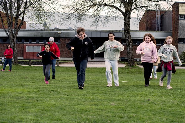 Fem barn kommer løpende mot kamera på en gressbane. De holder skjeer med poteter i hendene. 