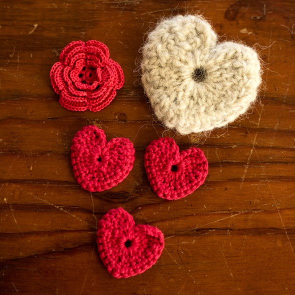 Tre røde og et hvitt heklet hjerte ligger på et trebord sammen med en heklet blomst