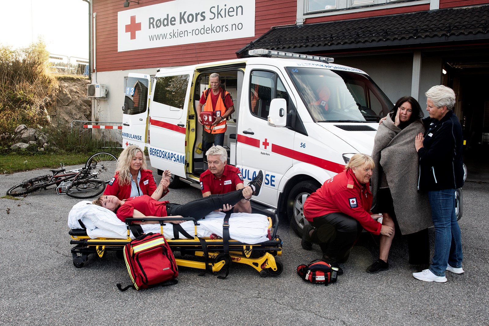 En beredskapsambulanse står foran et Røde Kors-hus og sju mennesker står rundt i forskjellige beredskapspositurer.
