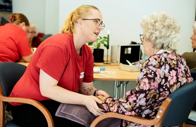 En ung dame sitter å snakker med en eldre dame mens hund holder hånden hennes.