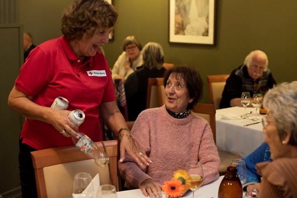 En frivillig står ved et bord med eldre damer og ler og prater med dem.