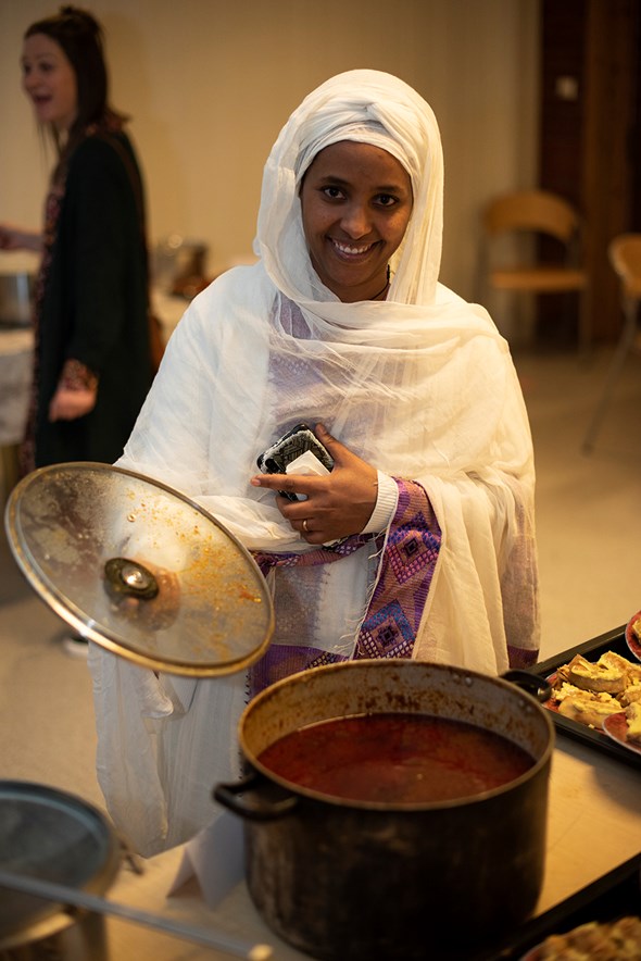 en dame står med sjal foran en gryte og smiler og viser fram maten hun har laget