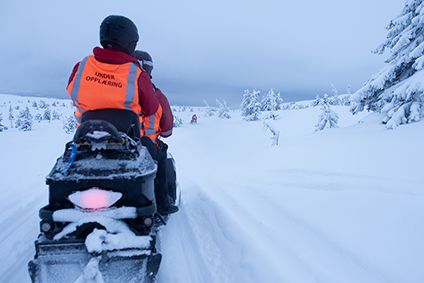 To scootere fotografert bakfra. På vidda i snøføyke. På ryggen til den bakerste står det under opplæring.