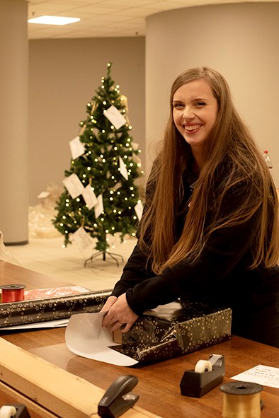 En smilende dame står foran et juletre og pakker inn julegave