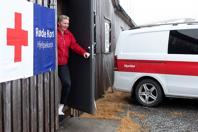 En dame kommer ut av en dør. Det står en Røde Kors-bil parkert utenfor og på veggen på hytta er det Røde Kors-logo