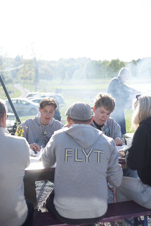 En gjeng mennesker sitter rundt et bord. Det er strålende sol og bildet dekkes av røyk fra grillen.