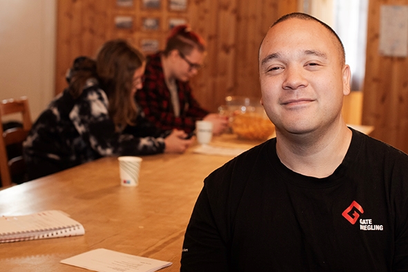 Bilde av en mann i forgrunn og to ungdommer som sitter ved et bord og ser på mobilene sine i bakgrunnen
