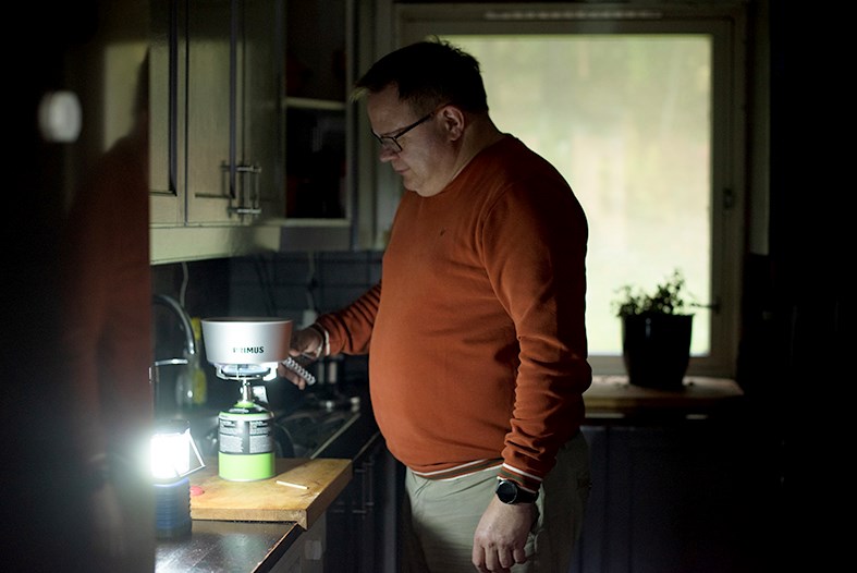 En mann står ved kjøkkenbenken kun opplyst av en liten nødlampe. Kjøkkenet er mørklagt og han koker vann på primus